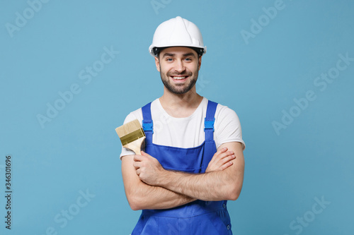 Smiling man in coveralls protective helmet hardhat hold paint brush isolated on blue wall background. Instruments accessories for renovation apartment room. Repair home concept. Holding hands crossed.