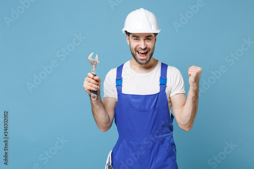 Excited man in coveralls protective helmet hardhat hold adjustable wrench isolated on blue background. Instruments accessories for renovation apartment room. Repair home concept. Doing winner gesture.