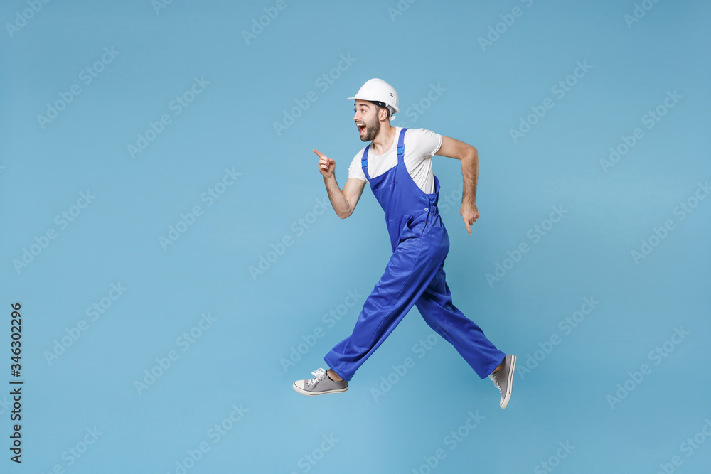 Side view of excited man in coveralls protective helmet hardhat isolated on blue background. Instruments accessories renovation apartment room. Repair home concept. Jumping, point index finger aside. Side view of excited man in coveralls protective helmet hardhat isolated on blue background. Instruments accessories renovation apartment room. Repair home concept. Jumping, point index finger aside.