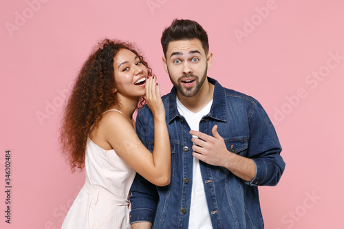 Amazed young couple two friends european guy african american girl in casual clothes posing isolated on pastel pink background. People lifestyle concept. Whispering secret behind hand, sharing news.