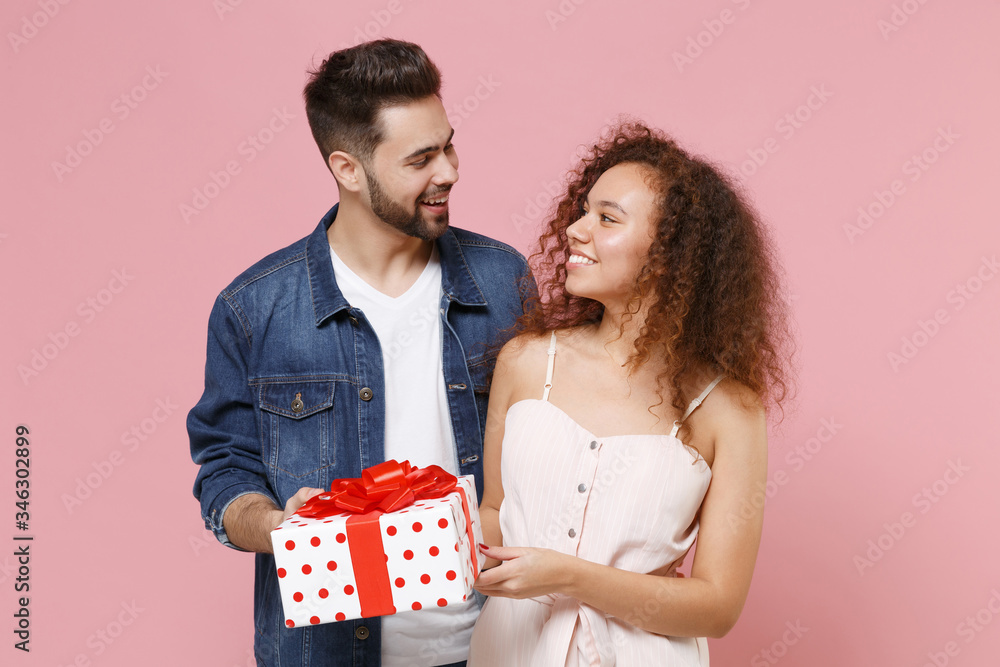 Smiling young couple friends european guy african american girl isolated on pastel pink background. Valentine's Day, Women's Day, birthday, holiday concept. Hold red present box with gift ribbon bow. Smiling young couple friends european guy african american girl isolated on pastel pink background. Valentine's Day, Women's Day, birthday, holiday concept. Hold red present box with gift ribbon bow.