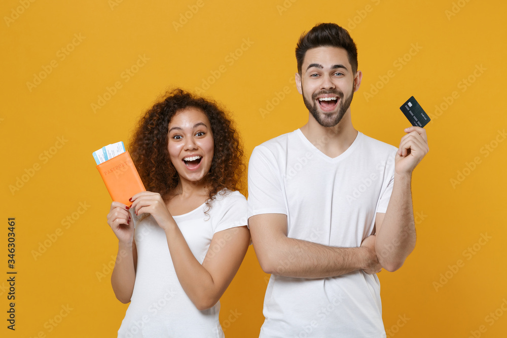 Excited young couple two friends european guy african american girl in white t-shirts posing isolated on yellow background in studio. People lifestyle concept. Hold credit bank card, passport tickets.