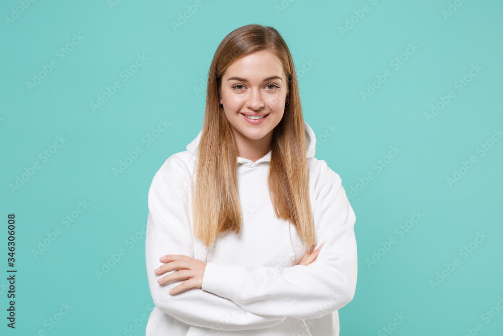Smiling young woman girl in casual white hoodie posing isolated on blue turquoise wall background studio portrait. People sincere emotions lifestyle concept. Mock up copy space. Holding hands crossed. Smiling young woman girl in casual white hoodie posing isolated on blue turquoise wall background studio portrait. People sincere emotions lifestyle concept. Mock up copy space. Holding hands crossed.