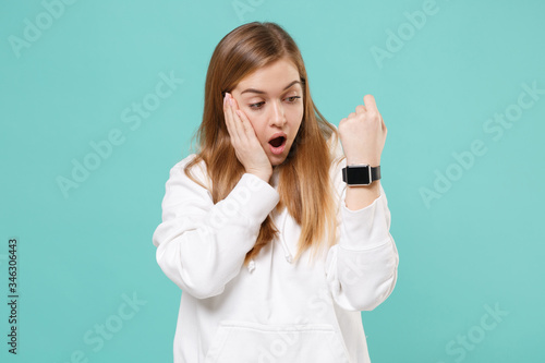 Shocked young woman in casual white hoodie posing isolated on blue turquoise background studio portrait. People lifestyle concept. Mock up copy space. Looking on smart watch on hand put hand on cheek.