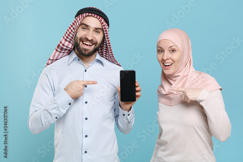 Funny couple friends arabian muslim man wonam in keffiyeh kafiya ring igal agal hijab clothes isolated on blue background. People religious lifestyle concept. Point on mobile phone with blank screen. photo