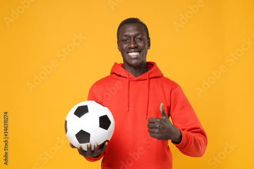 Smiling young african american man guy football player in red streetwear hoodie isolated on yellow wall background. Sport leisure lifestyle concept. Playing football hold soccer ball showing thumb up. © ViDi Studio