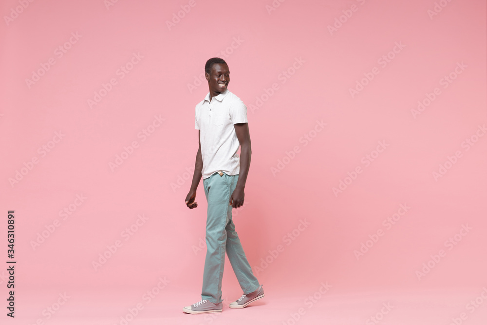 Side view of smiling young african american man guy in white polo shirt, turquoise trousers posing isolated on pastel pink wall background. People lifestyle concept. Mock up copy space. Looking aside. Side view of smiling young african american man guy in white polo shirt, turquoise trousers posing isolated on pastel pink wall background. People lifestyle concept. Mock up copy space. Looking aside.