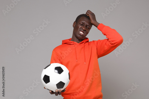 Displeased young african american man guy football player in orange streetwear hoodie isolated on grey background. Sport leisure lifestyle concept. Play football hold soccer ball put hand on head.