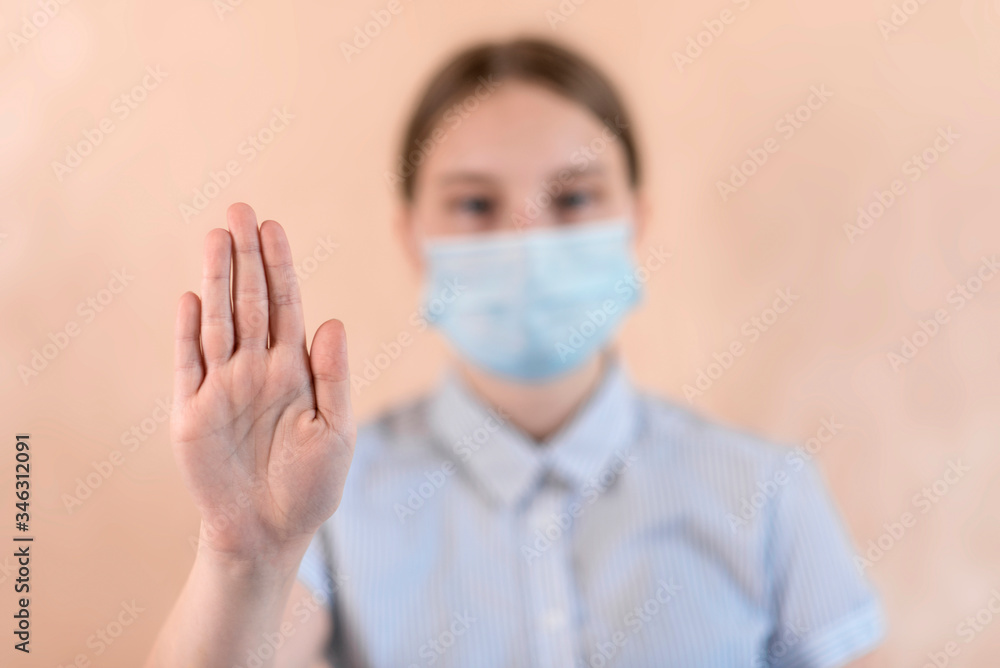 Teenager girl in protective medical mask covers her face, hand gesture stops attention and carefully virus. Flu infection is a disease and pandemic. Beige background.