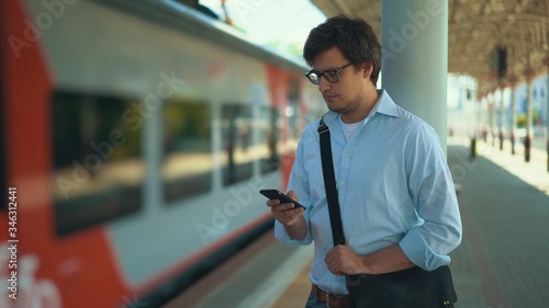 Young businessman wearing a blue shirt and glasses is texting while standing on the platform waiting for his train. Sunny summer day 