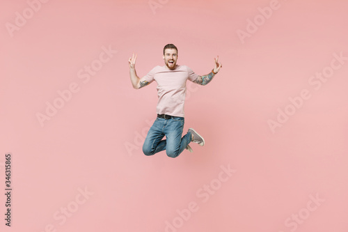 Excited young tattooed man guy in pastel casual t-shirt posing isolated on pink background studio portrait. People sincere emotions lifestyle concept. Mock up copy space. Jumping showing victory sign. © ViDi Studio