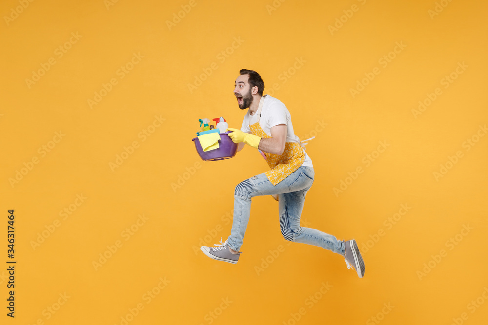Side view of crazy man househusband in apron rubber gloves hold basin detergent bottles washing cleansers doing housework isolated on yellow background. Housekeeping concept. Jumping like running.
