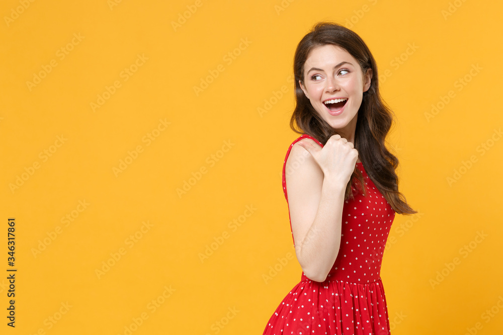 Side view of funny young brunette woman girl in red summer dress posing isolated on yellow background studio portrait. People sincere emotions lifestyle concept. Mock up copy space. Point thumb aside. Side view of funny young brunette woman girl in red summer dress posing isolated on yellow background studio portrait. People sincere emotions lifestyle concept. Mock up copy space. Point thumb aside.