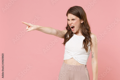 Irritated young brunette woman girl in light casual clothes posing isolated on pastel pink background. People lifestyle concept. Mock up copy space. Pointing index finger aside, screaming swearing. © ViDi Studio