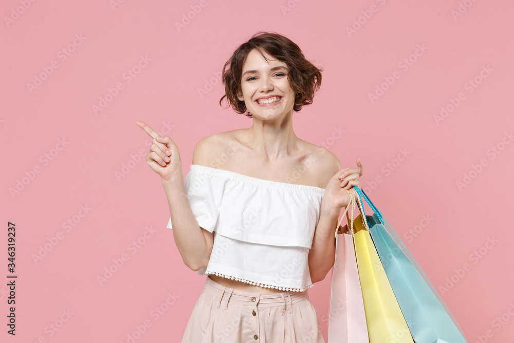 Smiling young woman girl in summer clothes hold package bag with purchases isolated on pink background in studio. Shopping discount sale concept. Mock up copy space. Pointing index finger aside up. Smiling young woman girl in summer clothes hold package bag with purchases isolated on pink background in studio. Shopping discount sale concept. Mock up copy space. Pointing index finger aside up.