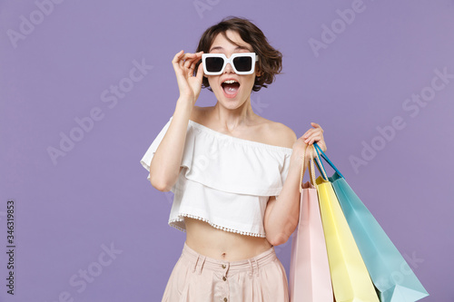 Shocked young woman girl in summer clothes eyeglasses hold package bag with purchases isolated on violet background in studio. Shopping discount sale concept. Mock up copy space. Keeping mouth open.