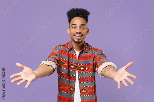 Excited young african american guy in casual colorful shirt posing isolated on violet background studio portrait. People emotions lifestyle concept. Mock up copy space. Stand with outstretched hands.