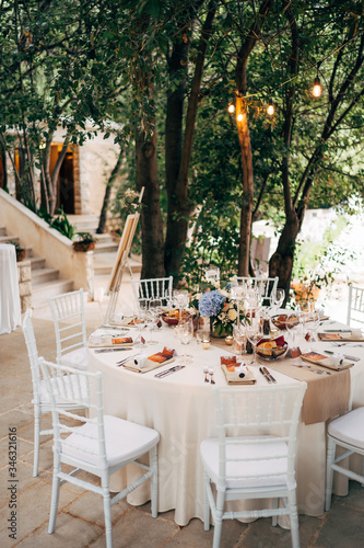 Wedding dinner table reception. Round table with white tablecloth with brown runner on table. Vienna nested in brown napkins, a bouquet of flowers is on the table. White Chiavari chairs with pillows. © Nadtochiy