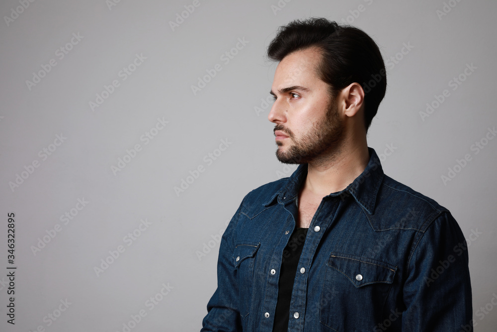 Side view of bearded man in the denim outfit which mixed with bright white t-shirt posing over white wall.