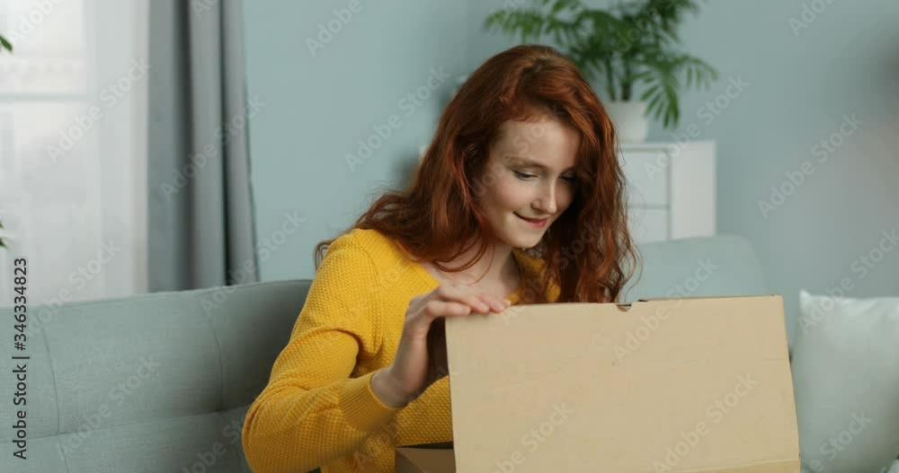 Happy young Caucasian woman with red hair opening carton parcel and smiling while sitting on sofa in living room. Cheerful red-haired girl taking out small boxes from cardboard box on couch at home. Happy young Caucasian woman with red hair opening carton parcel and smiling while sitting on sofa in living room. Cheerful red-haired girl taking out small boxes from cardboard box on couch at home.