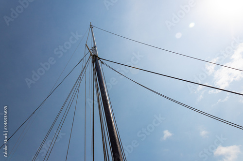 Mast of a vintage sailing vessel with numerous rigging lines coming from the top in many directions, against a bright blue sky with clouds, horizontal aspect