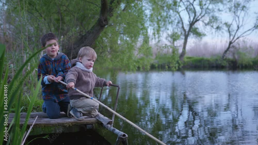 friendly children, cute boys sit on a wooden pier and fish using a fishing rod from river background of reeds friendly children, cute boys sit on a wooden pier and fish using a fishing rod from river background of reeds