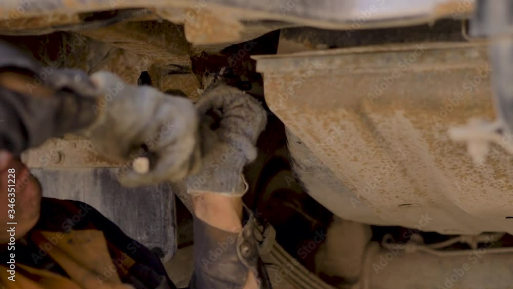 An Asian man is repairing the chassis of an old car lifted on a lift in a workshop, close-up. An Asian man is repairing the chassis of an old car lifted on a lift in a workshop, close-up.