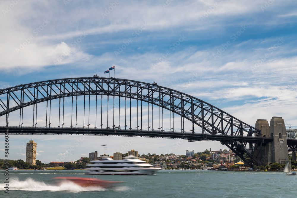 Fototapeta premium A ferry boat passing under the Sydney Harbour Bridge structure with cloudy blue sky above and city buildings in the background.