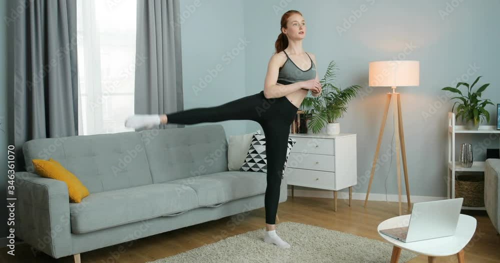 Young Caucasian woman working out and training her body in cozy living room during confinement. Pretty red-haired female exercising for legs muscles while lifting leg at home. Self-isolated sport. Young Caucasian woman working out and training her body in cozy living room during confinement. Pretty red-haired female exercising for legs muscles while lifting leg at home. Self-isolated sport.