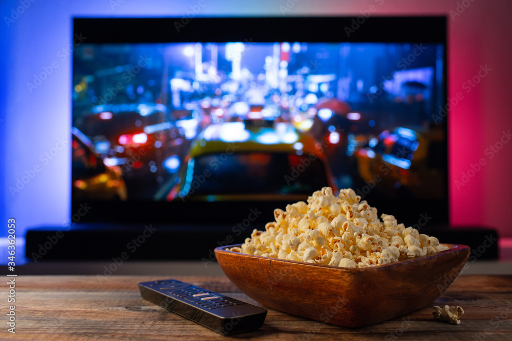 A wooden bowl of popcorn and remote control in the background the TV works. Evening cozy watching a movie or TV series at home - obrazy, fototapety, plakaty 