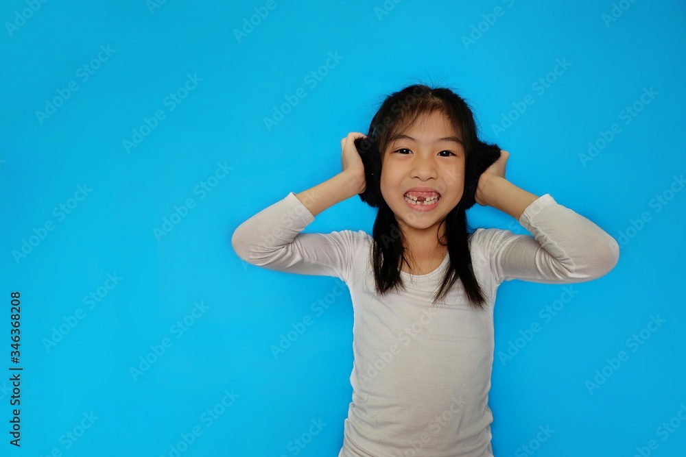 Fototapeta premium Cute młoda dziewczyna azjatyckich słuchanie muzyki przy użyciu jej czarnych puszystych słuchawek, uśmiechnięta, dobra zabawa. Zwykłe jasnoniebieskie tło.