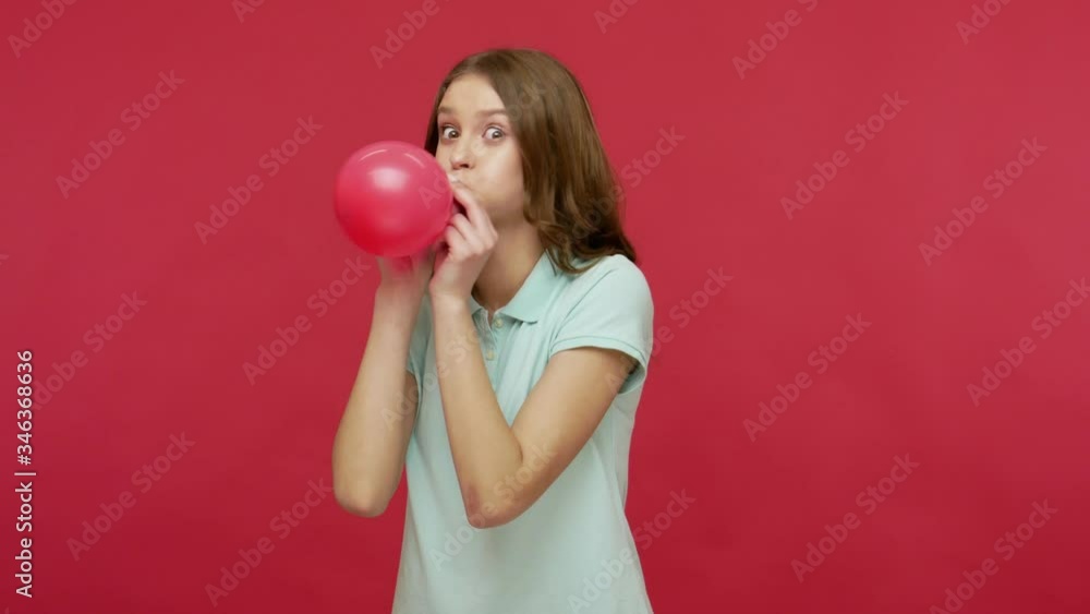 Funny happy young brunette woman in polo t-shirt inflating balloon with great effort, laughing and dancing, getting ready for party, celebrating birthday. indoor studio shot isolated on red background Funny happy young brunette woman in polo t-shirt inflating balloon with great effort, laughing and dancing, getting ready for party, celebrating birthday. indoor studio shot isolated on red background