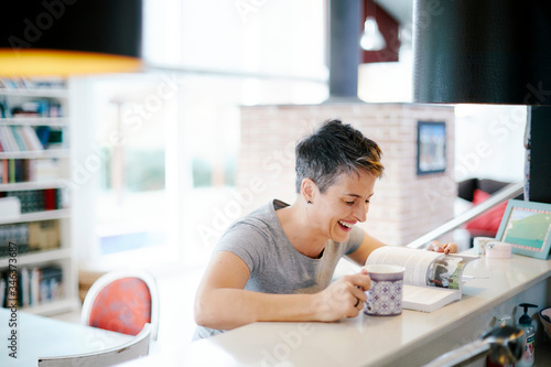 Short-haired woman with a cup and a book inside the house and laughing