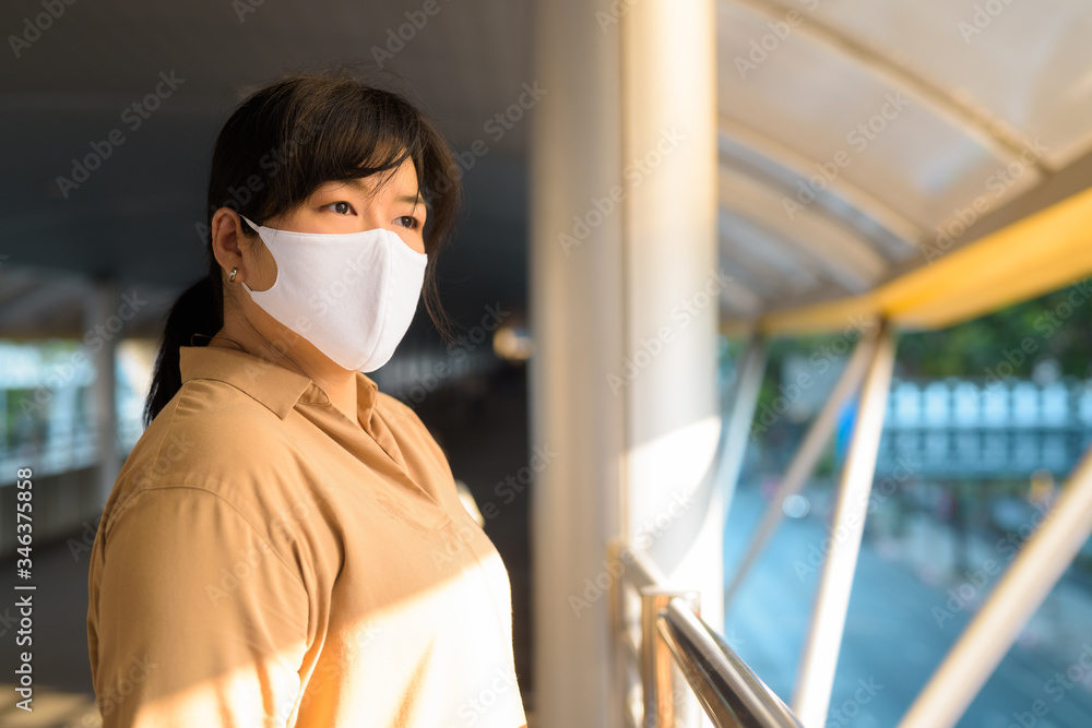 Overweight Asian woman with mask thinking at footbridge in the city
