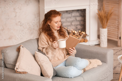 Beautiful young woman drinking hot cocoa and reading book at home