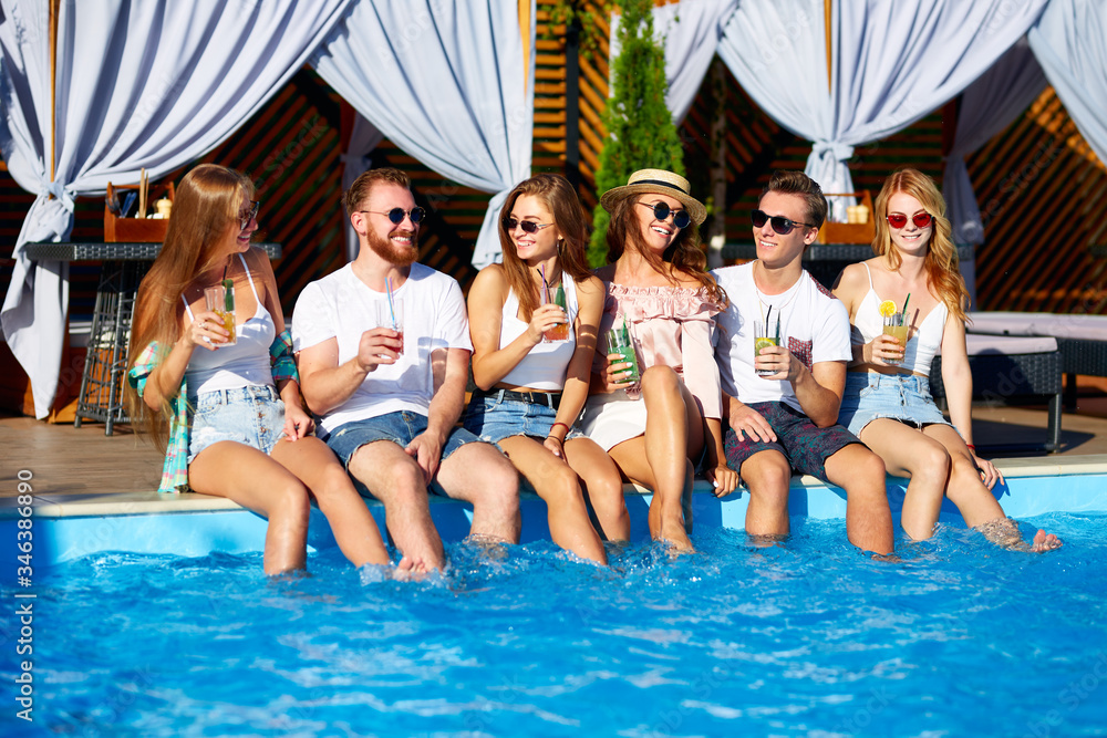 Group of friends having fun at poolside party clinking glasses with fresh cocktails sitting by swimming pool on sunny summer day. People toast drinking beverages at luxury villa on tropical vacation.