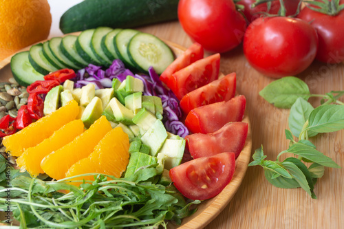 A buddha bowl of fresh summer healthy rainbow salad with cucumbers, tomatoes, arugula, basil, pumpkin seeds, avocado, red cabbage and chilli pepper on a wooden surface
