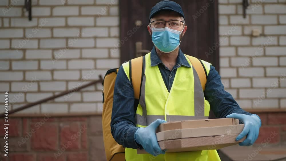 Delivery service worker with a yellow backpack in a medical mask and gloves holds cardboxes on the background of the house. Work during the coronavirus epidemic Delivery service worker with a yellow backpack in a medical mask and gloves holds cardboxes on the background of the house. Work during the coronavirus epidemic