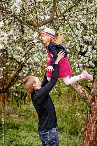 A young father plays with his daughter in a flowering garden. Throws up. Against the background of green grass and flowering trees.