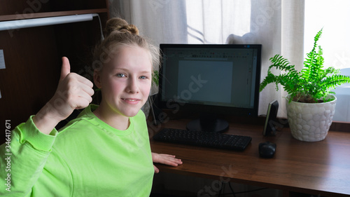 Teenager girl using desk computer for distance online learning in your room. Look at camera. Smiley face. Self-isolation. Thumbs up.