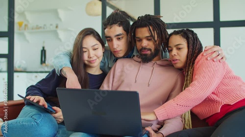 Group of joyful diverse multracial young people gathered on sofa, watching funny content online with laptop pc, chatting and laughing while enjoying leisure together in loft apartment. photo