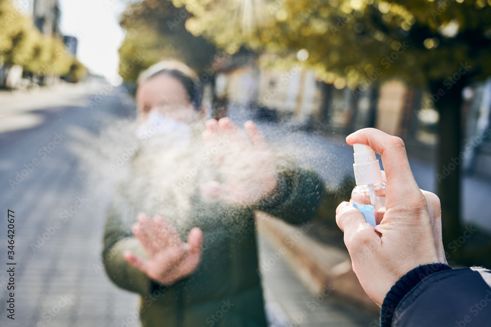 Selective focus of male's hand spraying alcohol based liquid to protect from coronavirus spreading. Woman in mask places her hands under spraying on background. Stop coronavirus infection concept Selective focus of male's hand spraying alcohol based liquid to protect from coronavirus spreading. Woman in mask places her hands under spraying on background. Stop coronavirus infection concept
