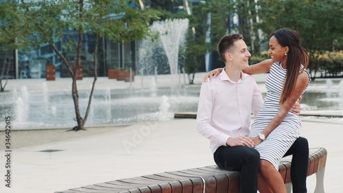 Romantic multiracial couple having a date: woman whispering something in boyfriend's ear. They sitting on a bench, embracing and kissing.
