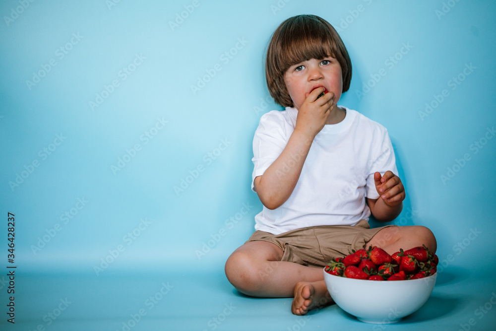 Toddler boy sitting with bowl of strawberries on the blue background Toddler boy sitting with bowl of strawberries on the blue background