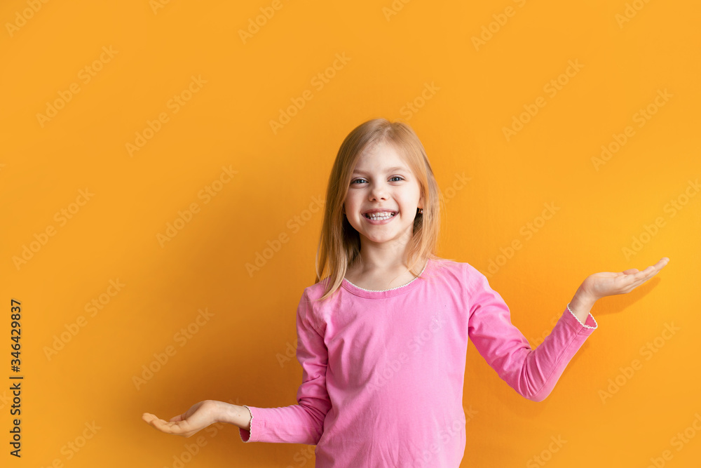 Cute baby on an orange background, 6-8 years old, a girl in pink clothes smiles, shows the background, points to a specific place Cute baby on an orange background, 6-8 years old, a girl in pink clothes smiles, shows the background, points to a specific place