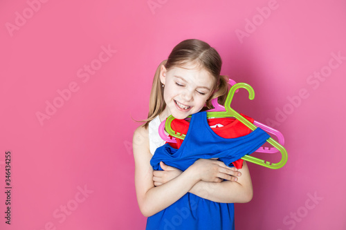Baby on a pink background, little girl holds two dresses of different colors, rejoices in buying two for the price of one