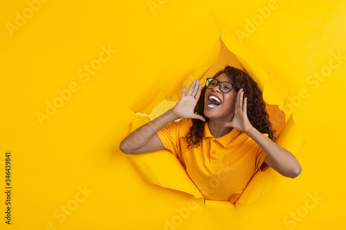 Astonished shouting. Cheerful african-american young woman in torn yellow paper background, emotional, expressive. Breaking on, breakthrought. Concept of human emotions, facial expression, sales, ad.