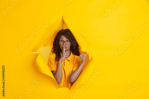 Secretly pointing. Cheerful african-american young woman in torn yellow paper background, emotional, expressive. Breaking on, breakthrought. Concept of human emotions, facial expression, sales, ad. © master1305
