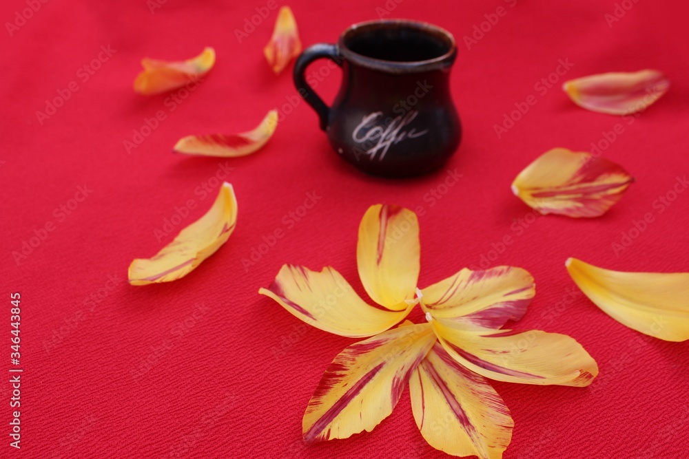 A small brown cup with the inscription Coffee isolated on a red background surrounded by petals. Cup of coffee on a background of tulip petals.