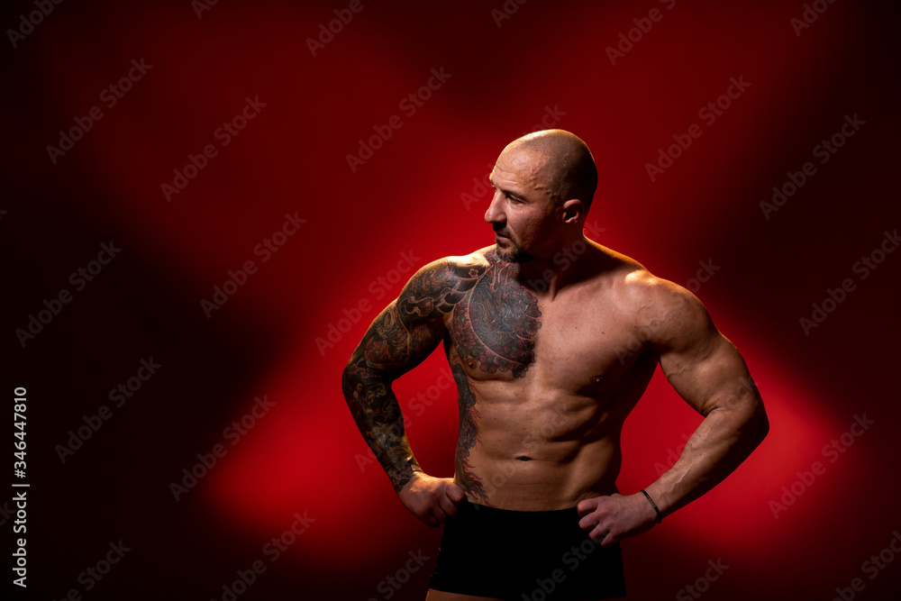 A sports man with a tattoo posing on red background. Fitness trainer with naked torso, standing in the studio. Bodybuilder looking away A sports man with a tattoo posing on red background. Fitness trainer with naked torso, standing in the studio. Bodybuilder looking away